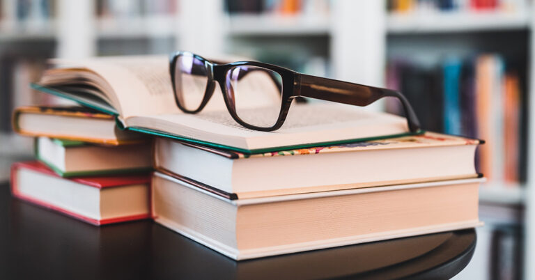 Ein Stapel Bücher liegt auf einem dunklen Tisch, obendrauf eine Brille mit schwarzem Rand. Im Hintergrund befindet sich verschwommen ein Bücherregal.