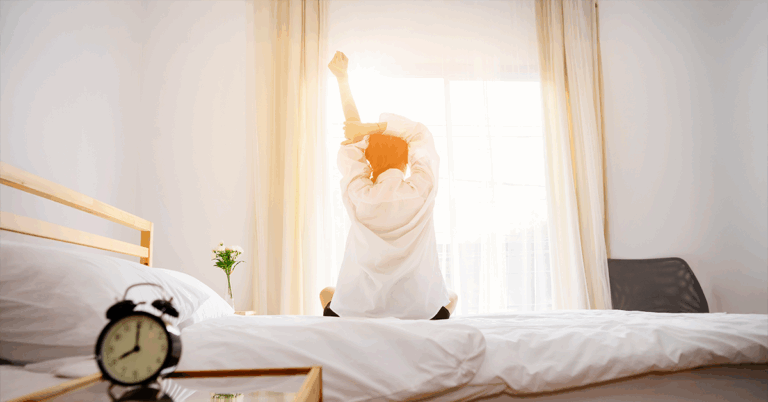 Eine Frau sitzt auf ihrem Bett in einem hellen, sonnigen Schlafzimmer und streckt sich, um ihre Morgenroutine zu beginnen.