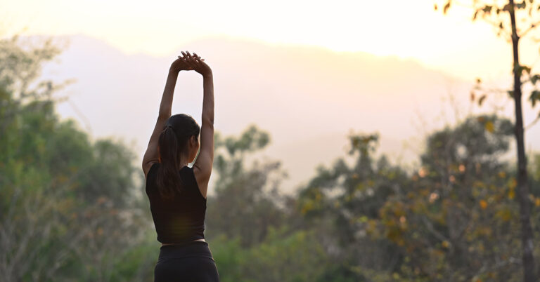 Weibliche Person macht Dehnungsübungen im Freien, das Licht ist weich und warm wie bei einem Sonnenaufgang. Auch Stretching ist Outdoor-Training und kann viel für das eigene Wohlbefinden tun.
