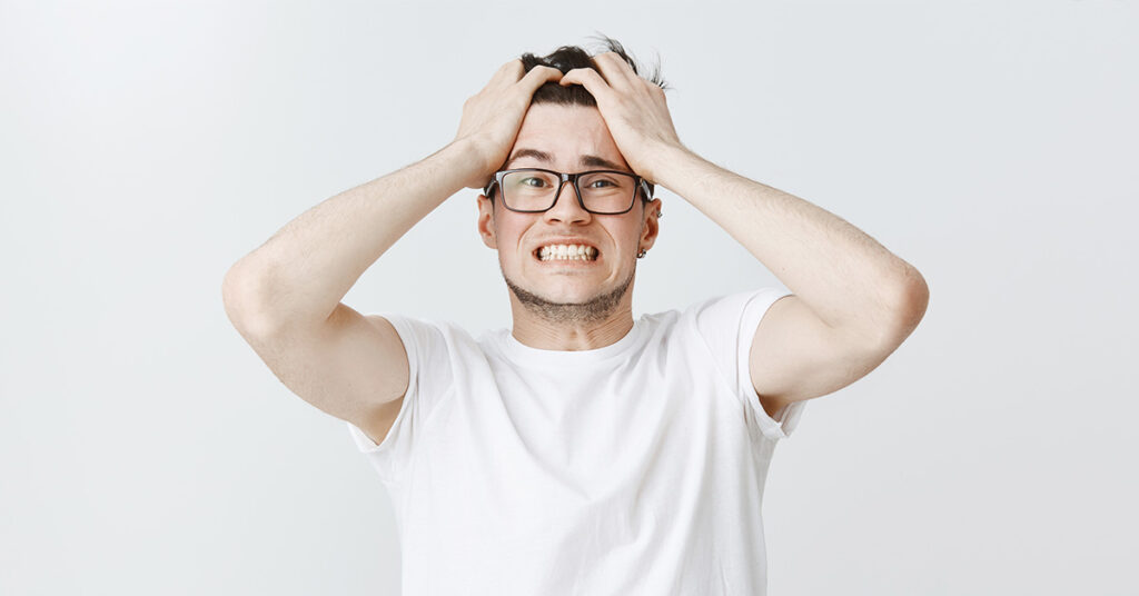 Junger Mann steht vor einer weißen Wand, greift sich mit beiden Händen an den Kopf und beißt nervös die Zähne zusammen. Er blickt besorgt in die Kamera und scheint an Overthinking zu verzweifeln.