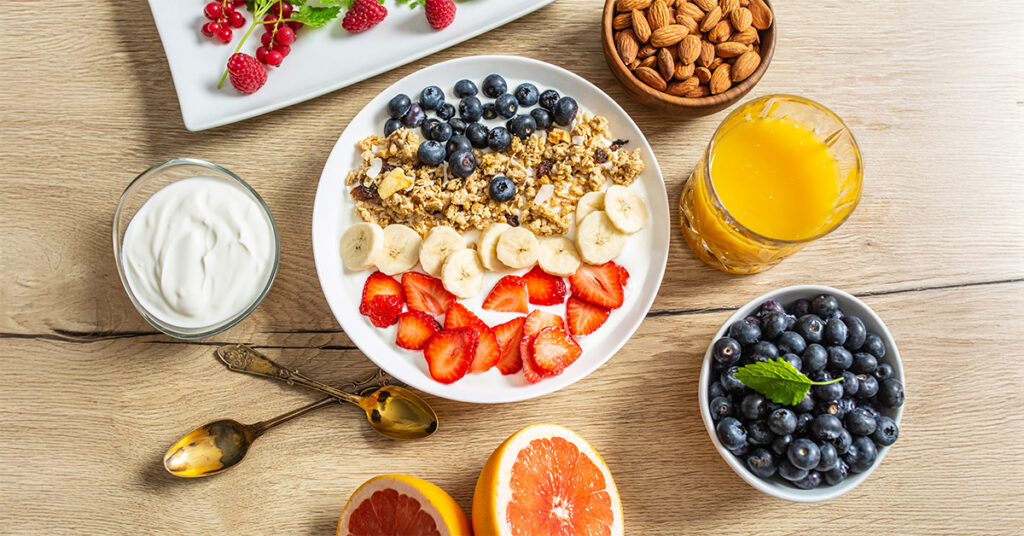 Gesunde Nahrungsmittel auf einem Holztisch: Eine Schüssel mit augeschnittenen Früchten und Joghurt, eine Schale Nüsse, ein Glas Saft und weitere.