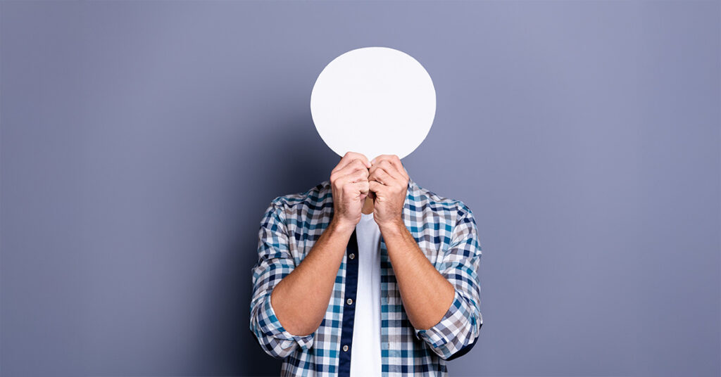 Introvertierte Person: Mann im karierten Hemd steht vor einer grau-lila Wand und hält sich ein rundes weißes Schild vors Gesicht.