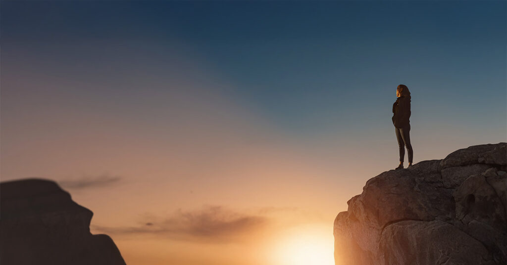 Silhouette einer Frau, die während des Sonnenaufgangs auf einem Berg steht, sie wirkt einsam.