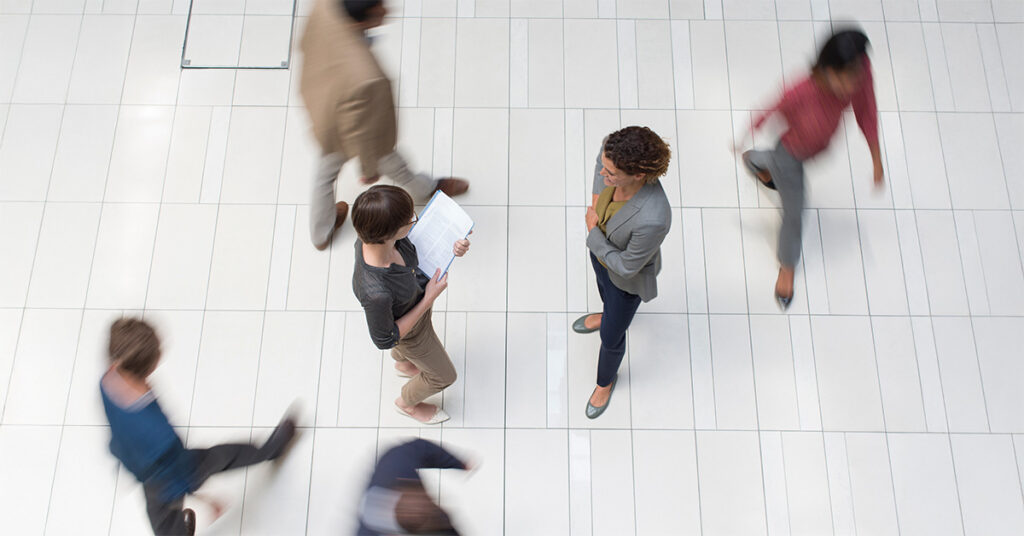 Aufnahme von schräg oben: zwei Frauen unterhalten sich, andere Personen laufen (verschwommen dargestellt) an ihnen vorbei. Das Bild steht symbolisch für Energie und Umfeld.