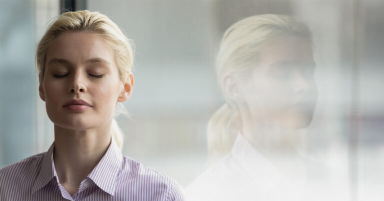 Mental Decluttering: Eine blonde Frau im gestreiften Hemd steht mit geschlossenen Augen und entspanntem Gesichtsausdruck vor einer Scheibe, in der sie sich seitlich spiegelt.