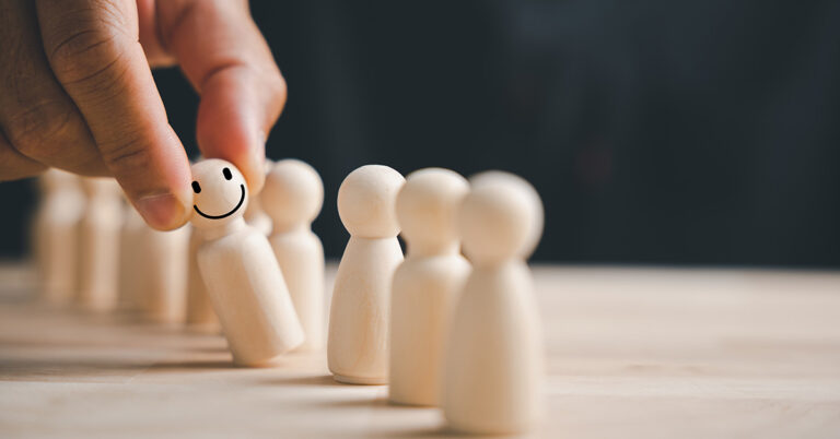 Eine Hand nimmt eine Holzfigur aus einer Reihe anderer Figuren, sie trägt ein Smiley-Gesicht.