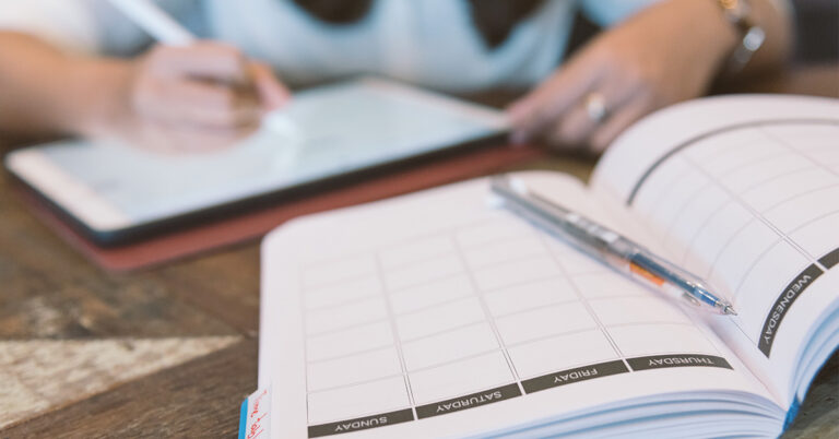 Eine Person stzt an einem Tisch, vor sich ein Tablet und ein Kalender, und plant ihre Woche.