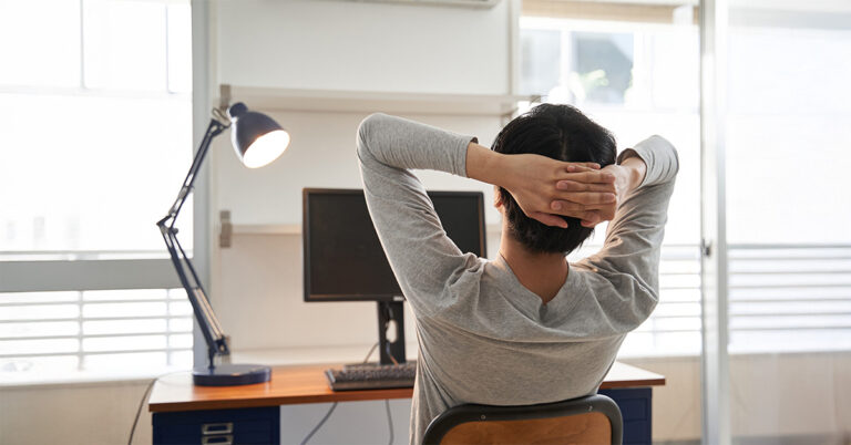 Eine Person sitzt mit hinter dem Kopf verschränkten Armen vor einem ausgeschalteten Laptop